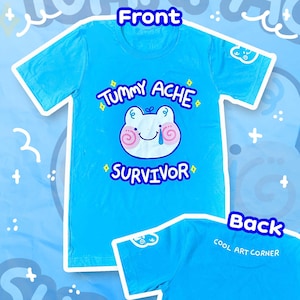 May include: A light blue t-shirt with the words "Tummy Ache Survivor" and a cartoon frog design. The back of the shirt has the text "COOL ART CORNER".