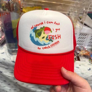 May include: Red and white trucker hat with a graphic of a fish jumping out of the water. The hat has the text "of course i cum fast i got FISH to catch!!!!!!!!!" on the front.