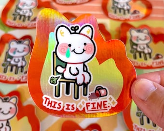 Puede incluir: Pegatina holográfica con un oso de dibujos animados blanco con una lágrima verde, sentado en una silla, rodeado de llamas. El texto "THIS IS FINE" está debajo. La pegatina tiene un borde rojo y naranja en forma de llama.