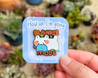 May include: A square enamel pin with a cartoon character holding a blue blanket. The pin has the text "Hold up, I'm going BLANKIE MODE" in gold and white. The background is light blue with white diamond shapes.