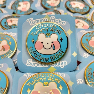 Puede incluir: Un pin de esmalte dorado con fondo azul y un conejito de dibujos animados blanco con mejillas rosadas. El pin dice "Official Tummy Ache Survivor Badge" en letras doradas.