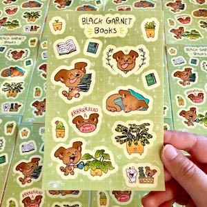 Puede incluir: Una hoja de pegatinas con ilustraciones de perros leyendo libros, haciendo jardinería y durmiendo. Las pegatinas están sobre un fondo verde con el texto "Black Garnet Books" en la parte superior.