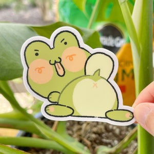 May include: A green frog sticker with a cartoonish style. The frog has a big smile and is sticking its tongue out. The frog is sitting on its hind legs with its front legs crossed. The sticker is on a green plant.