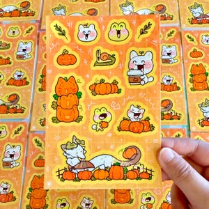 Puede incluir: Una hoja de pegatinas con dibujos animados de animales y calabazas. Las pegatinas están sobre un fondo amarillo con un patrón de cuadrícula. Los animales incluyen gatos, ranas y caracoles. Las calabazas son de color naranja y tienen una variedad de expresiones.