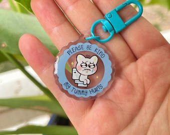 Puede incluir: Un llavero azul y blanco con un gato de dibujos animados en un inodoro. El texto en el llavero dice "Por favor, sé amable, me duele la barriga."