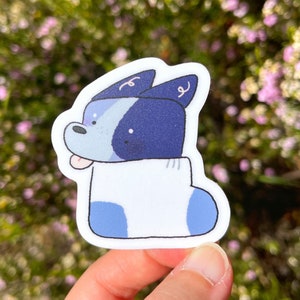 May include: A blue and white cartoon dog sticker with a white sock on its paw. The dog has a blue nose and black eyes.