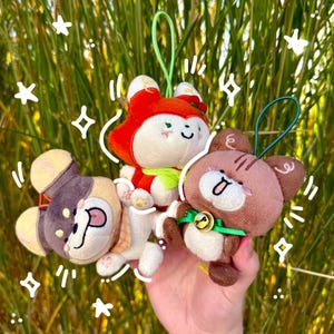 May include: Three plush animal keychains in the image. The keychains are a fox, a bear, and a dog. The fox is red and white, the bear is brown, and the dog is tan and purple. Each keychain has a loop for hanging.