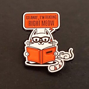 Peut inclure: Une broche en émail blanc représentant un chat portant des lunettes noires et lisant un livre orange. Une bulle au-dessus du chat contient le texte GO AWAY, I'M READING RIGHT MEOW. Une petite souris blanche est située à droite du chat.