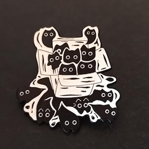 Peut inclure: Épingle en émail noir et blanc représentant une boîte débordant d'illustrations de chats noirs. L'épingle a un design unique avec plusieurs visages de chats et des formes fantomatiques, idéale pour les amoureux des chats.