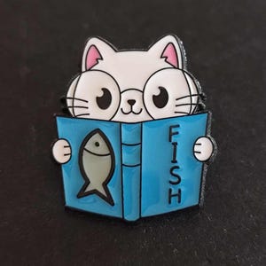 Peut inclure: Un chat blanc portant des lunettes lit un livre bleu avec le mot "FISH" sur la couverture et une illustration de poisson à l'intérieur.