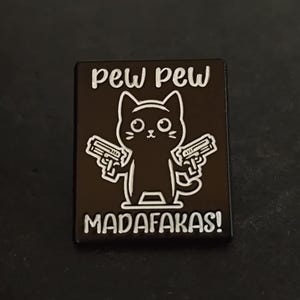 Pode incluir: Pin de esmalte preto com um gato de desenho animado segurando duas armas. O pin tem as palavras "pew pew" acima do gato e "MADAF*KAS!" abaixo, tudo em letras brancas. O gato tem olhos grandes.