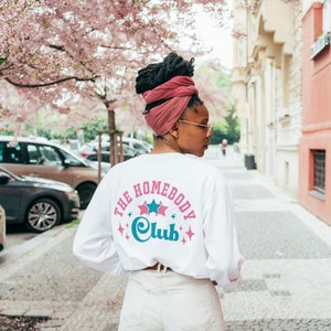 May include: A white long-sleeved shirt with the text "The Homebody Club" in pink and blue lettering with stars around the text. The person is wearing a pink turban.