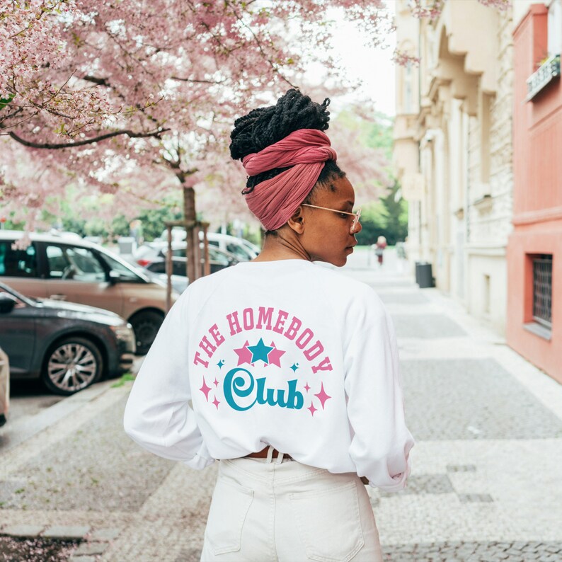 May include: A white long-sleeved shirt with the text "The Homebody Club" in pink and blue lettering with stars around the text. The person is wearing a pink turban.