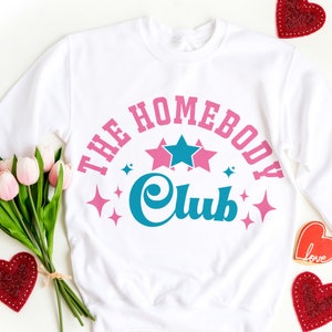 May include: White sweatshirt with a pink and blue graphic design that says "The Homebody Club" with stars and sparkles.