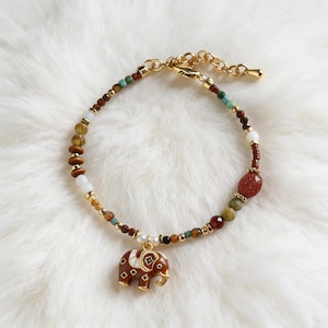 Puede incluir: Pulsera dorada con un dije de elefante marrón. La pulsera presenta una variedad de cuentas coloridas, incluyendo marrón, verde y blanco. El dije de elefante es marrón con detalles blancos. Sobre una superficie blanca y esponjosa.