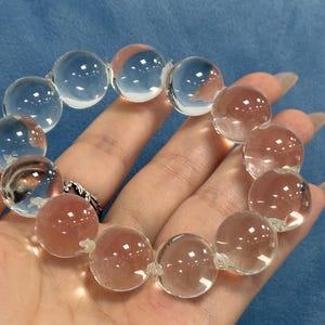 Pode incluir: Uma pulseira de contas transparentes e quartzo rosa. A pulseira apresenta grandes contas redondas enfiadas juntas. As contas são transparentes com uma ligeira tonalidade rosa. A pulseira é segurada em uma mão sobre um fundo azul.
