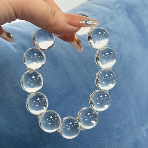 Puede incluir: Pulsera de cuentas redondas y transparentes. Las cuentas están ensartadas para formar una forma circular. La pulsera se sostiene sobre un fondo azul. Las cuentas parecen ser de vidrio o un material transparente similar.