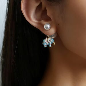 May include: A pair of earrings featuring a light blue elephant charm with red and pink accents. The elephant has gold-colored tusks and is suspended from a small gold-colored hoop. A white pearl stud is visible behind the ear.