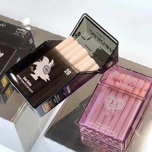 May include: Two cigarette cases, one black and one pink, are displayed. The black case, labeled "Son Goku," holds cigarettes. The pink case, labeled "Pink," also contains cigarettes. Both cases have graphic designs and text.