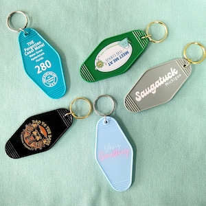 May include: Five different motel keychains with various designs. The keychains are all in the shape of a motel key. The keychains are blue, green, gray, black, and light blue. The keychains have different text on them, including "The Forgotten Coast Motel", "Meet Me At The Lodge", "Saugatuck Michigan", "Bad To The Bone", and "Very Something".