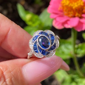 May include: A silver-toned ring featuring a central oval-cut deep blue sapphire surrounded by a floral-shaped halo of smaller square-cut blue sapphires and tiny round clear stones, all set within a detailed metal frame with milgrain edges.