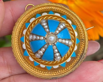 May include: A gold and turquoise enamel brooch with a central pearl and smaller pearls around the edge. The brooch is circular and has a detailed design.