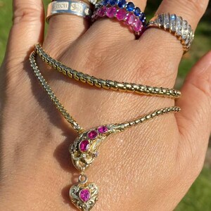 May include: A gold-toned snake necklace with pink gemstones and a heart pendant. Several rings with clear and pink gemstones are also visible. The jewelry is displayed on a hand against a blurred green background.