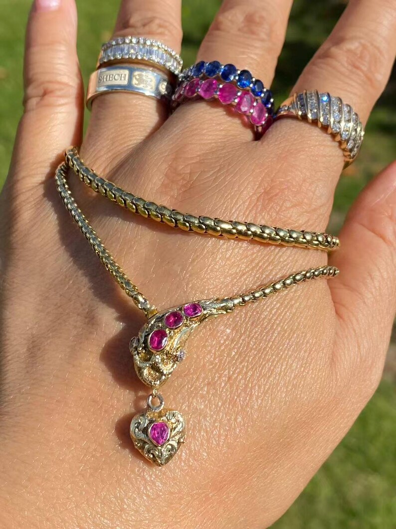 May include: A gold-toned snake necklace with pink gemstones and a heart pendant. Several rings with clear and pink gemstones are also visible. The jewelry is displayed on a hand against a blurred green background.