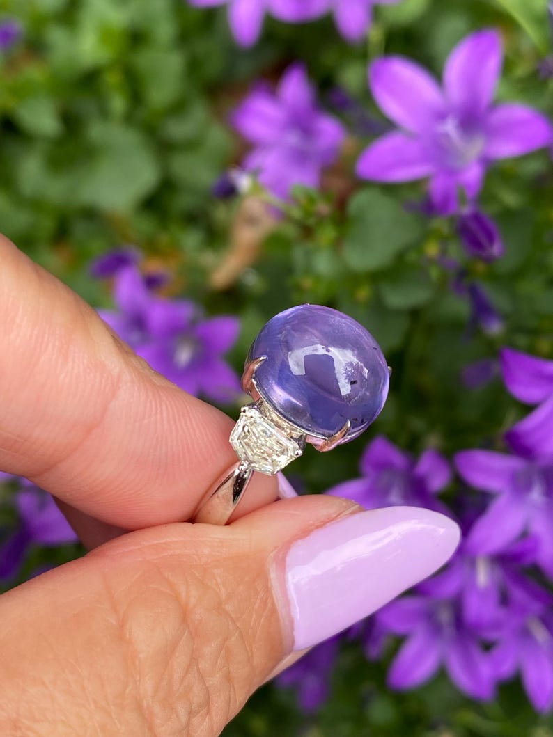 May include: A silver ring featuring a large, round, translucent purple gemstone, flanked by two smaller, clear, rectangular stones. The ring is held against a backdrop of blurred purple flowers and green foliage.
