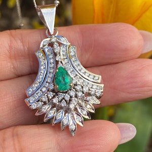 Pode incluir: Um colar com pingente de prata com uma esmeralda em forma de pêra rodeada por pequenos diamantes brilhantes. O pingente tem um design ornamental, de inspiração floral, com uma corrente delicada.