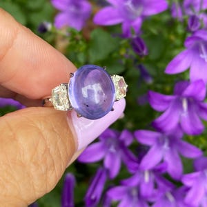 May include: A gold ring featuring a large, oval, lavender-colored gemstone flanked by two smaller, rectangular, clear gemstones. The ring is held against a backdrop of vibrant purple flowers, creating a striking contrast of colors and textures. The ring's design is elegant and eye-catching.