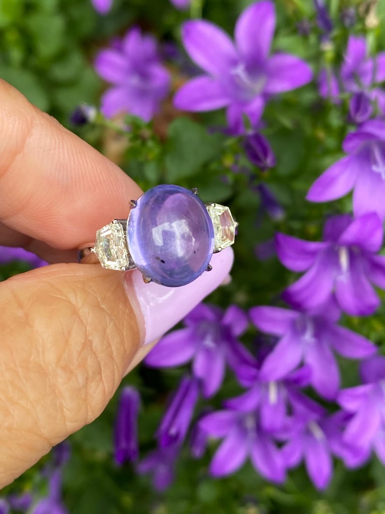 May include: A gold ring featuring a large, oval, lavender-colored gemstone flanked by two smaller, rectangular, clear gemstones. The ring is held against a backdrop of vibrant purple flowers, creating a striking contrast of colors and textures. The ring's design is elegant and eye-catching.