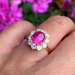 May include: A gold ring with a large, oval-cut, bright pink gemstone at its centre, encircled by smaller, clear gemstones. The ring has a thin gold band. The background is blurred, with pink flowers.