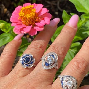 May include: Three silver-toned rings featuring blue and clear stones. The left ring has a central oval blue stone surrounded by smaller blue and clear stones. The middle ring features a round clear stone with blue accents. The right ring has a central clear stone with blue and clear stone detailing.