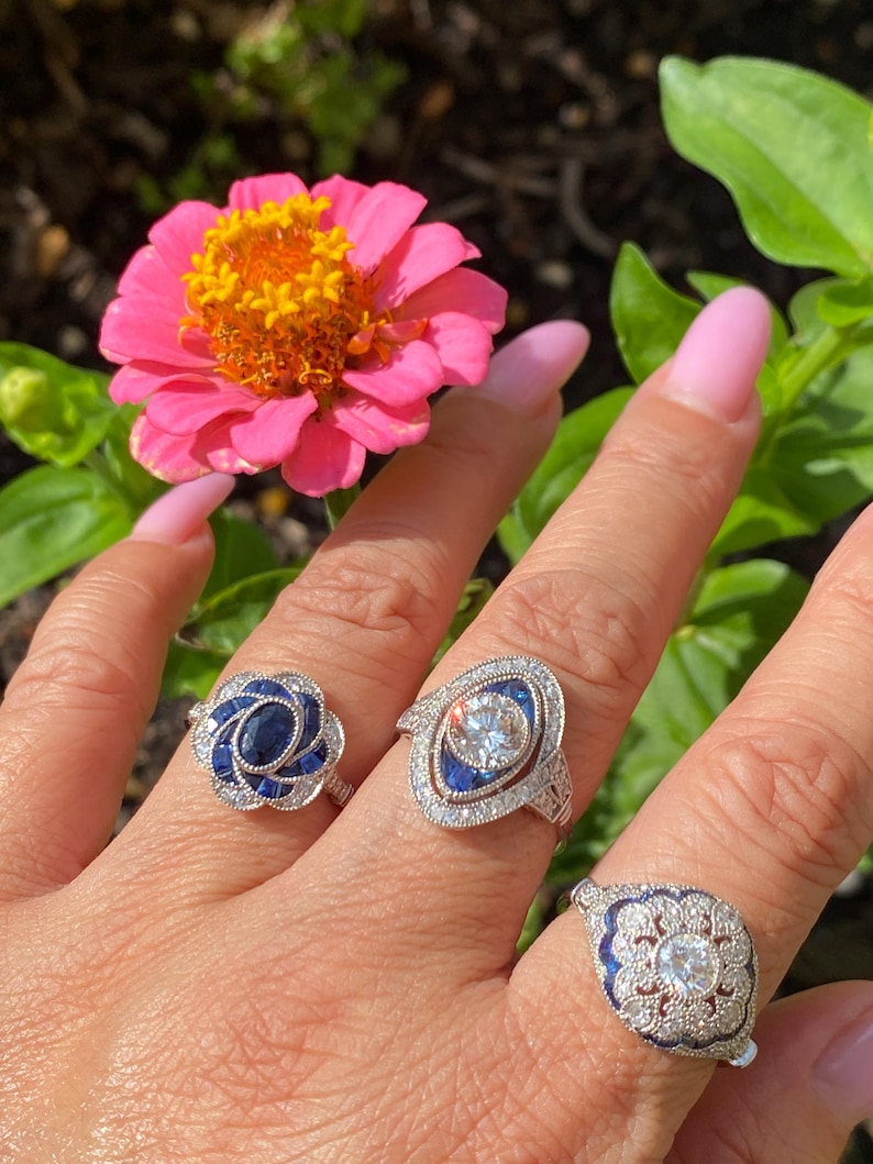 May include: Three silver-toned rings featuring blue and clear stones. The left ring has a central oval blue stone surrounded by smaller blue and clear stones. The middle ring features a round clear stone with blue accents. The right ring has a central clear stone with blue and clear stone detailing.