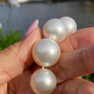 Pode incluir: Um colar de p&eacute;rolas brancas grandes. As p&eacute;rolas s&atilde;o redondas e t&ecirc;m uma superf&iacute;cie lisa e reflexiva. Est&atilde;o enfiadas com um fio branco fino. O fundo est&aacute; desfocado, com toques de verde e azul.