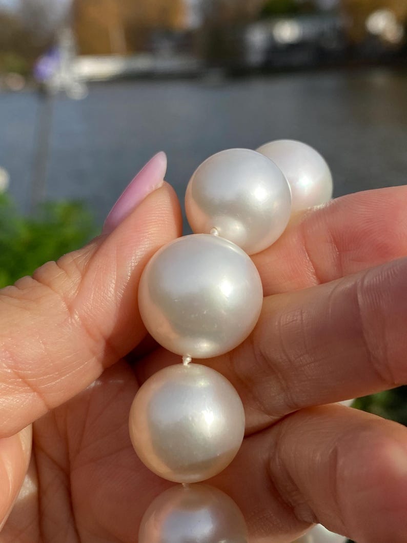 Pode incluir: Um colar de p&eacute;rolas brancas grandes. As p&eacute;rolas s&atilde;o redondas e t&ecirc;m uma superf&iacute;cie lisa e reflexiva. Est&atilde;o enfiadas com um fio branco fino. O fundo est&aacute; desfocado, com toques de verde e azul.