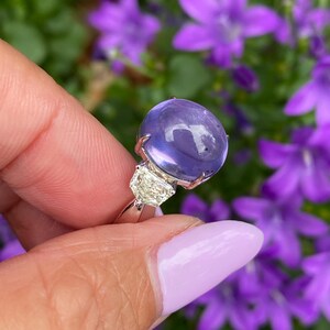 May include: A silver ring featuring a large, round, translucent purple gemstone and two smaller, clear, rectangular gemstones. The ring is held against a backdrop of blurred purple flowers and green foliage.