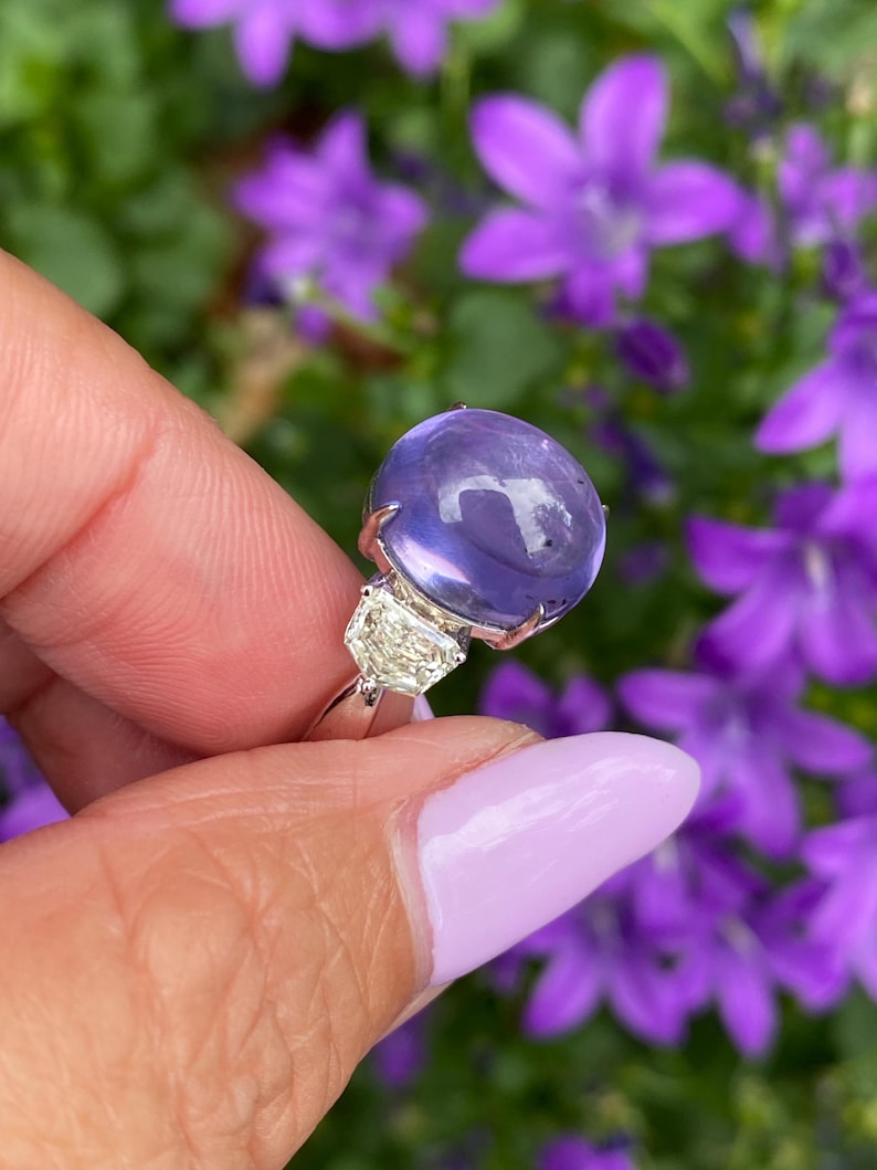 May include: A silver ring featuring a large, round, translucent purple gemstone and two smaller, clear, rectangular gemstones. The ring is held against a backdrop of blurred purple flowers and green foliage.