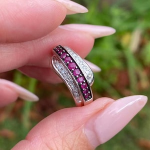 May include: A silver ring with a curved design, featuring a row of small, round, deep red gemstones and a row of smaller, clear gemstones. The ring is held up against a blurred green background.