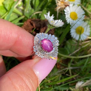 May include: A ring featuring a large, oval, purplish-pink gemstone with a star-like effect, surrounded by a halo of smaller, clear gemstones. The ring is set in a silver or white metal setting. The background includes green grass and white flowers.