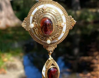 May include: A gold pendant with a large, oval, red gemstone at the center, surrounded by a white enamel border with gold accents and small pearls. A smaller, teardrop-shaped pendant with a matching red gemstone hangs below.