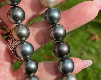 Op de afbeelding: Een ketting van donkergrijze en lichtbruine parels. De ronde parels hebben een hoge glans en zijn geregen met een zilveren sluiting. De ketting wordt tegen een vage groene achtergrond gehouden.