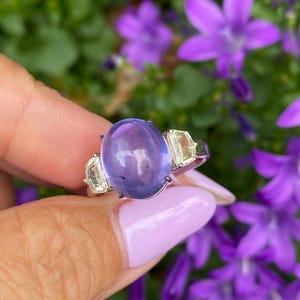 May include: A silver ring featuring a large, oval, lavender-colored gemstone flanked by two smaller, rectangular, light yellow gemstones. The ring is set against a backdrop of green foliage and purple flowers.