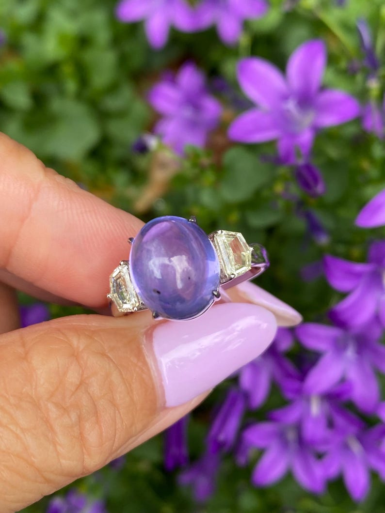 May include: A silver ring featuring a large, oval, lavender-colored gemstone flanked by two smaller, rectangular, light yellow gemstones. The ring is set against a backdrop of green foliage and purple flowers.