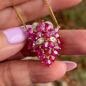 May include: A gold necklace featuring a cluster pendant of oval-cut, vibrant red gemstones, possibly rubies, accented with small, clear gemstones. The pendant is suspended from a delicate gold chain. The jewelry is held up against a blurred background.