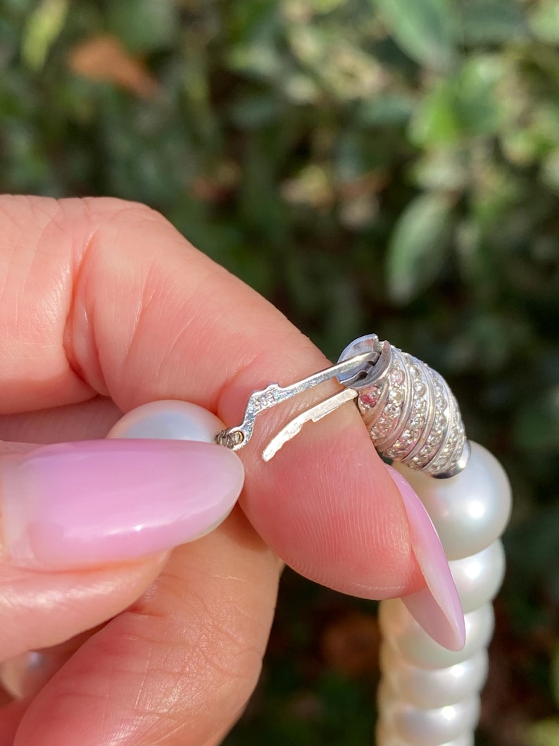 Pode incluir: Close-up de um colar de p&eacute;rolas com fecho prateado. O fecho &eacute; adornado com pequenas pedras brilhantes. As p&eacute;rolas s&atilde;o grandes e brancas, enfiadas para formar uma pe&ccedil;a de joalharia cl&aacute;ssica. O fundo &eacute; verde desfocado.