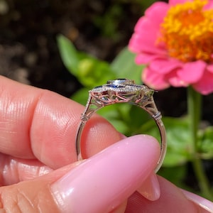 May include: A silver-toned ring featuring a central dark blue gemstone surrounded by a halo of smaller blue stones and clear accent stones. The metal band has an intricate openwork gallery design with milgrain detailing along the edges.