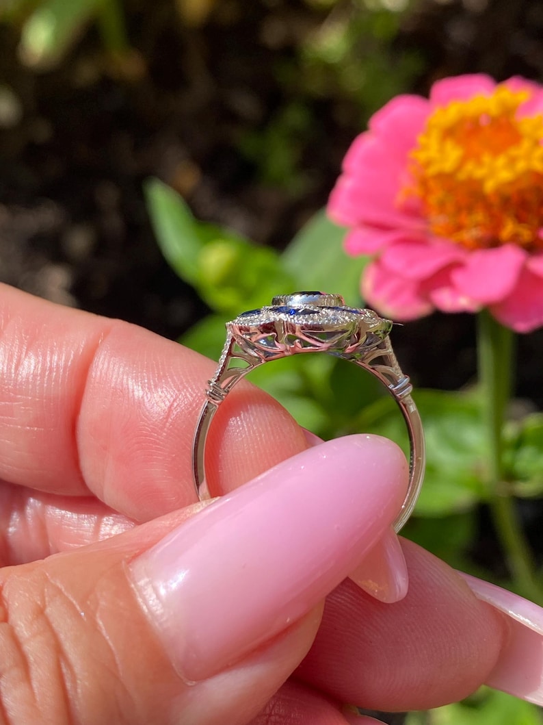 May include: A silver-toned ring featuring a central dark blue gemstone surrounded by a halo of smaller blue stones and clear accent stones. The metal band has an intricate openwork gallery design with milgrain detailing along the edges.