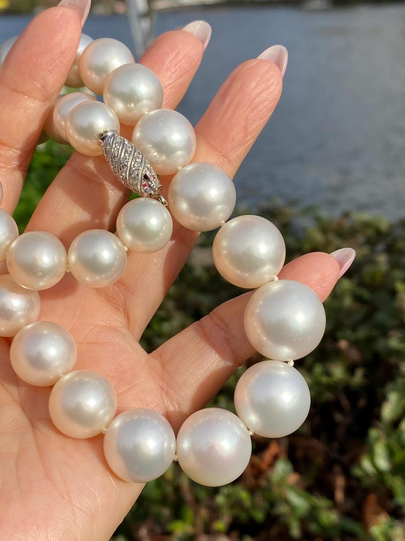 Pode incluir: Um colar de p&eacute;rolas brancas grandes e lustrosas. As p&eacute;rolas s&atilde;o redondas e de tamanho uniforme, com um alto brilho. O colar &eacute; preso com um fecho prateado, adornado com pequenas pedras transparentes e uma &uacute;nica pedra vermelha. As p&eacute;rolas est&atilde;o sendo seguradas.